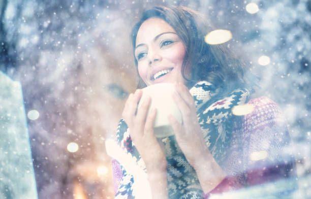Closeup of woman having a cup of tea or coffee on snowy day. She's sitting in a cafe and looking through window. Wearing knitted cardigan and scarf and enjoying winter in warm and cosy place. Low angle shot. Visible reflection in the window.