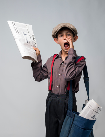 Little Newsboy Holding Newspapers And Shouting  To sell.The newspaper is in right hand and he is using his left hand for shouting.He is wearing a brown shirt red suspenders and black pans.His bag is full of newspapers.The image was shot with Hasselblad H4D on gray background in vertical composition.