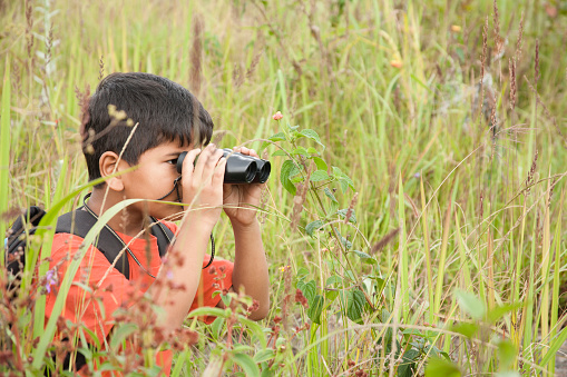 Cute little boy enjoys exploring the outdoor world using binoculars in a meadow or open grassy field.  The child wears a serious expression as he looks off into the distance to discover nature.  Science, education themes.  Elementary aged.  Latin, Indian, Asian descent child.   Tall grasses in foreground. 