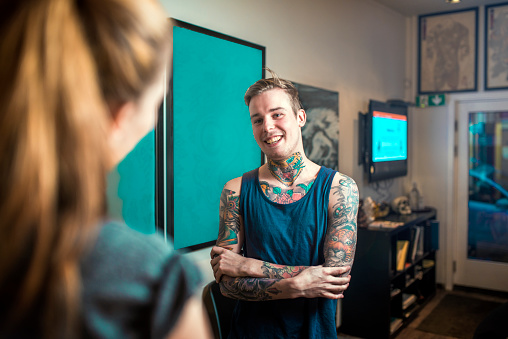 A photo of happy tattoo expert talking to customer. Tattooed man is attending female client. Both are in studio.