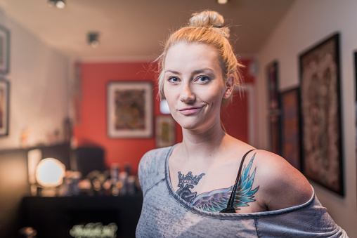 A photo of fashionable tattooed woman in studio. Portrait of confident female tattoo artist at workplace. Hipster is with tattoo on her chest.