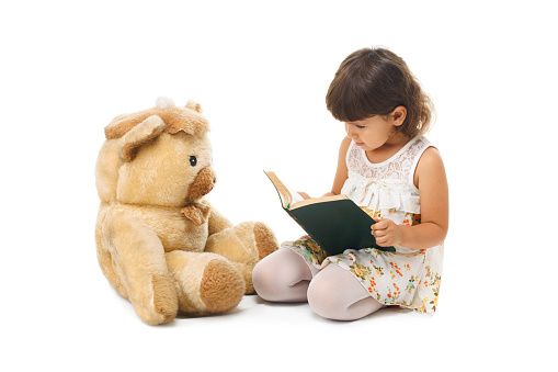 Little girl reading book  to her teddy bear isolated on white background