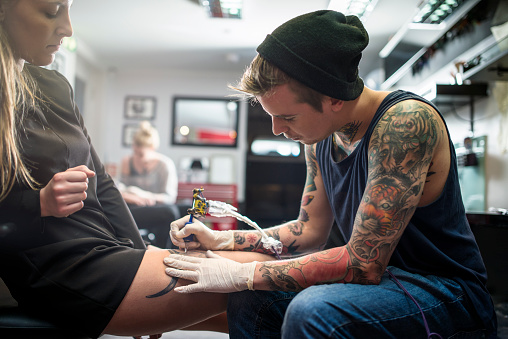 A photo of expert tattooing female customer's lap. Tattoo artist is working in studio. Woman is looking at man making tattoo on her leg.