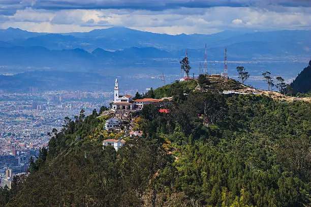The peak of Monserrate and beyond, as viewed from the neighbouring peak of Guadalupe on the Cordillera Oriental, in the capital city of Bogota in the South American country of Colombia.  The peak is about 500 metres above the capital city, which itself is over 2500  metres or 8450 feet,  above mean sea level on the Eastern Andes.  It is Sunday afternoon and a lot of people can be seen in front of the church on the peak. The distance however is too great to recognise anyone.The style of architecture of the church is typical Spanish colonial. Photo shot in the afternoon sunlight; horizontal format. Copy space.