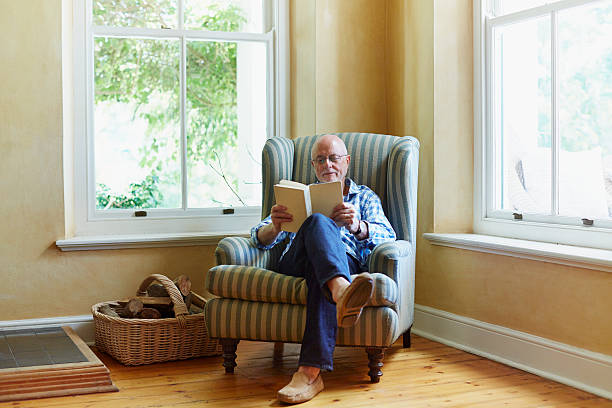 senior man reading book at home - armchair stock-fotos und bilder