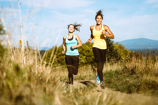 Two women run on a trail in a rural mountain area, smiles on their faces as they enjoy the exercise and warm afternoon. Depicting trail running, healthy lifestyle, friendship, and exercise in the great outdoors.  Horizontal image with copy space.