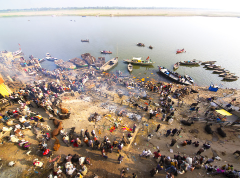 Varanasi, India - January 5, 2014: One of burning Ghats in centre of Varanasi with river Ganges in background. A lot of people standing on the cremation grounds while burning death person’s smoke rise from the fire into the air.