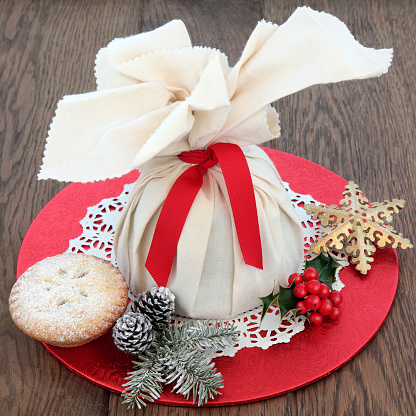 Christmas mince pies with pudding in a muslin bag, holly, winter greenery and snowflake bauble over oak background.