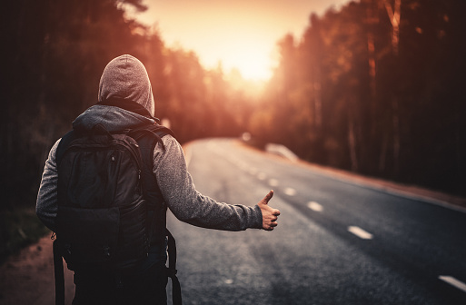 Hitchhiking traveler with backpack trying to stop the car on road in the forest at sunset. Stock photo.