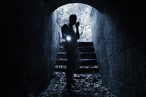 Young man with radio set and flashlight enters the stone tunnel and looks in the dark, monochrome photo