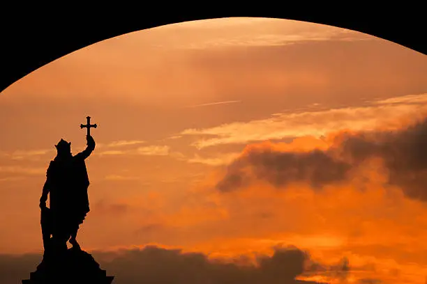 King Pelayo statue silhouetted under stone arch and sunset background. 