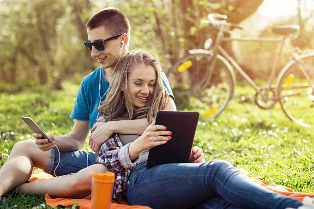 Teenage couple on a picnic Teenage couple hugging each other on a picnic in the nature. The boy is listening to music and wearing blue t-shirt, and the girl is looking at pictures on digital tablet and dressed in chequered shirt. In the background is a bicycle and spring meadow bathed in evening spring sunlight. teenager couple child blond hair stock pictures, royalty-free photos & images