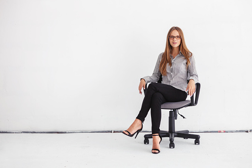 Portrait of young beautiful business woman sitting on chair against white wall.