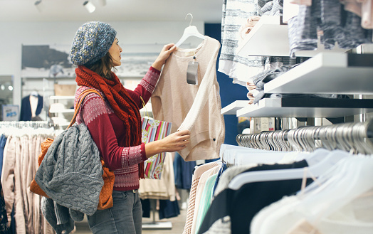 Closeup of smiling blond woman choosing clothes at department store  in local supermarket. She's holding a beige blouse and looking at it. The woman is wearing gray cap, red sweater and scarf. Side view.