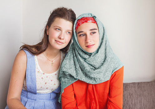 friendship of the religions concept: muslim and christian girl sitting on a background of a white wall