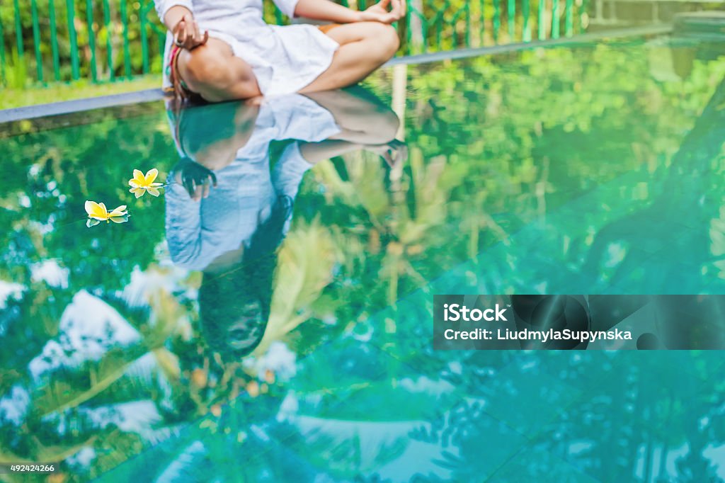 Woman meditating at pool side. Woman sitting in "lotus" pose on an edge of tropical swimming pool. Reflection in the water (faceless) 2015 Stock Photo