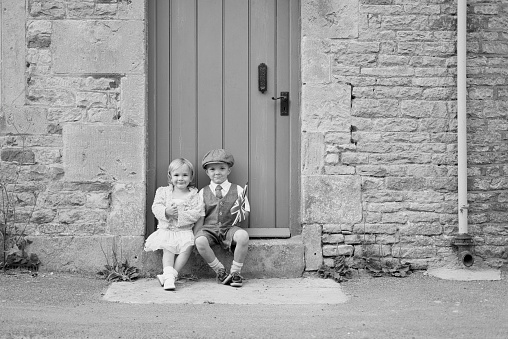 A young girl in vintage dress sits with a boy in his dapper duds on the step of an old English door. They are celebrating their English heritage by waving the national flag of the United Kingdom.