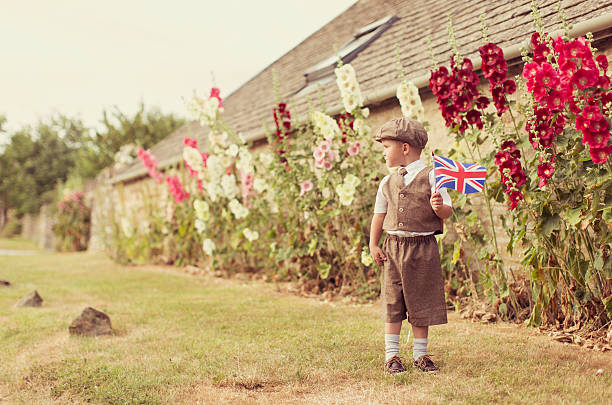 A young British boy in vintage clothing is celebrating his English heritage by waving the national flag of the United Kingdom. He is standing in front of holly hocks. Vintage toned.