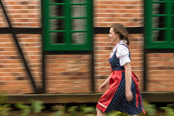 Woman in dirndl with bacon in hand in front of an alpine meadow with cows and the sign \