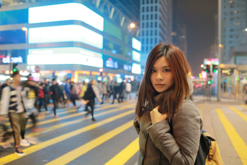 Beautiful young girl stand out and watching in after work at night in hong kong, lost in city , busy crowd and yellow zebra crossing blurred background
