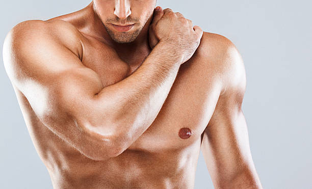 Muscular male upper body. Closeup of unrecognizable muscular man with one hand on opposite shoulder, looking down. He's standing shirtless in front of gray background. There's a bit of oil on his skin for better muscle definition. Torso stock pictures, royalty-free photos & images