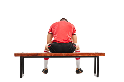 Rear view studio shot of a sad male football player sitting on a wooden bench and looking down isolated on white background