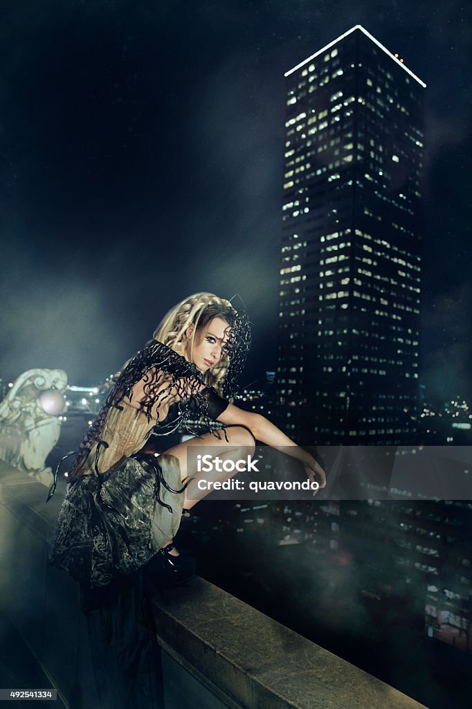 High Fashion Model Sitting on Ledge of Skyscrapper High fashion model sitting on ledge of a high rise building at night as if she's a gargoyle. Haute Couture Stock Photo