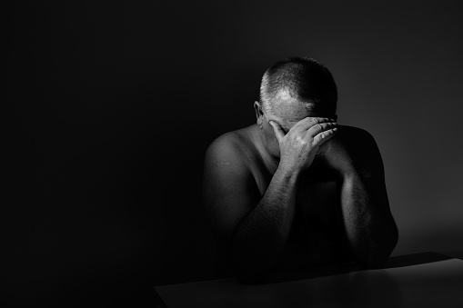 Dramatic black and white close up portrait of aged man sitting with hands on his face against wall - depression concept