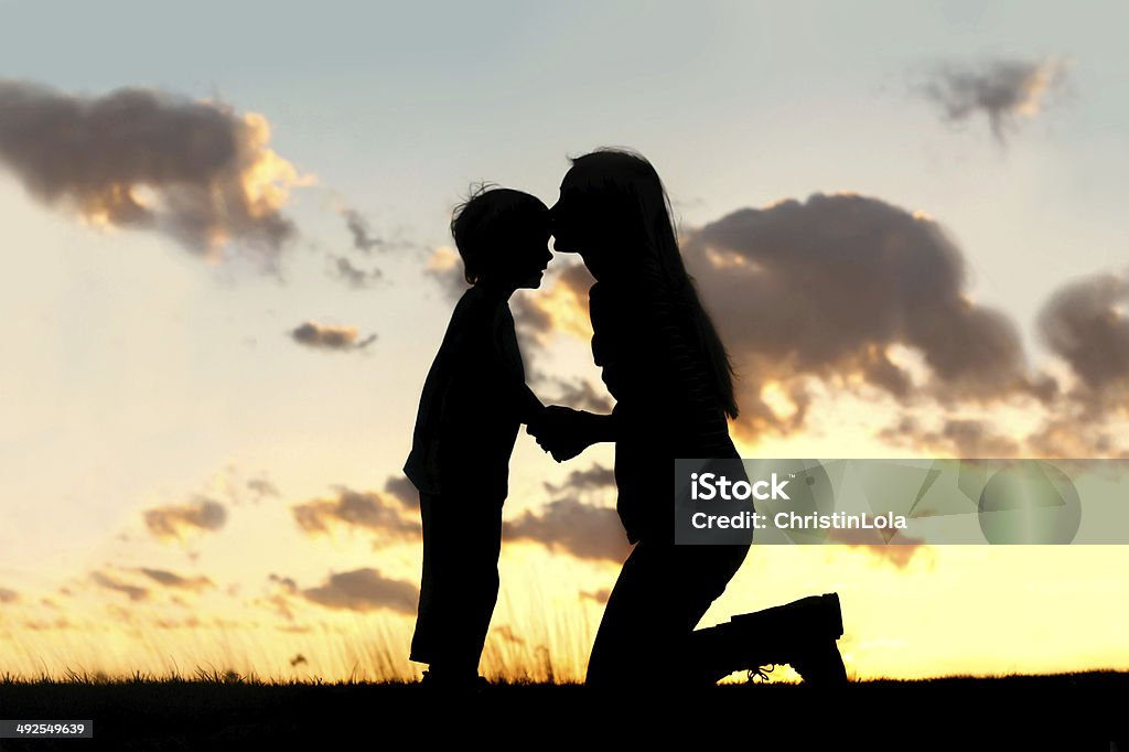 Mother Lovingly Kissing Little Child at Sunset Silhouette of a young mother lovingly kissing her little child on the forehead, outside isolated in front of a sunset in the sky. Mother Stock Photo