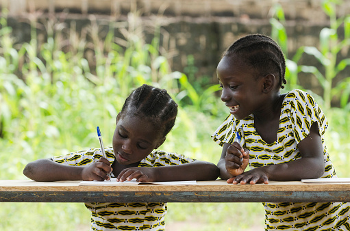 Schoolgirls repeating their lesson sitting in their desk at school in Bamako, Mali. Beautiful educational symbol. One girl's writing, the other one's slightly smiling.