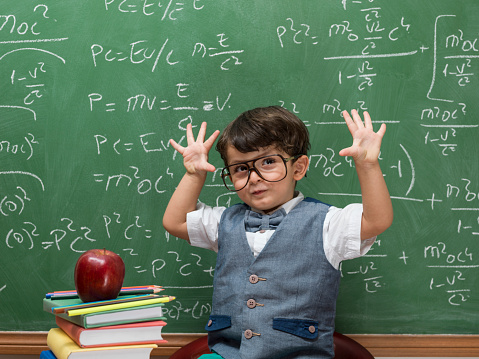 Little boy posing before blackboard with mathematic formulas written on.He is wearing horn rimmed glasses.Both hands raised for counting.Red apple placed on stack of books in front of model.The image was shot with a medium format camera Hasselblad in studio.