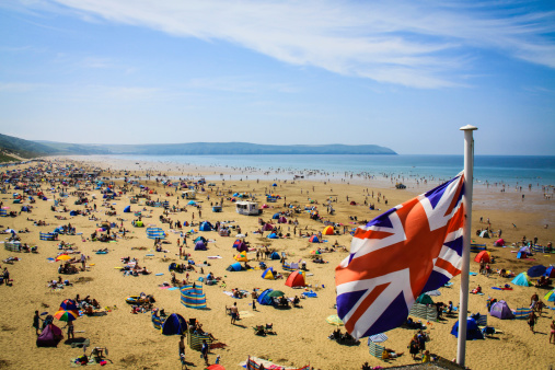 A VIEW OF WOOLACOMBE BEACH ON A BRITISH SUMMERS DAY, WITH A PROMINENT UNION JACK FLAG AND A BEACH FULL OF HOLIDAY MAKERS. BLUE SKIES AND A BLUE SEA.