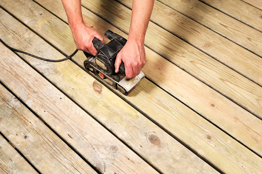 A worker is using a belt sander to sand and recondition weathered deck boards in preparation for staining.