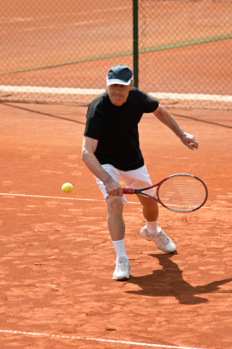 Senior serving playing tennis on clay court.