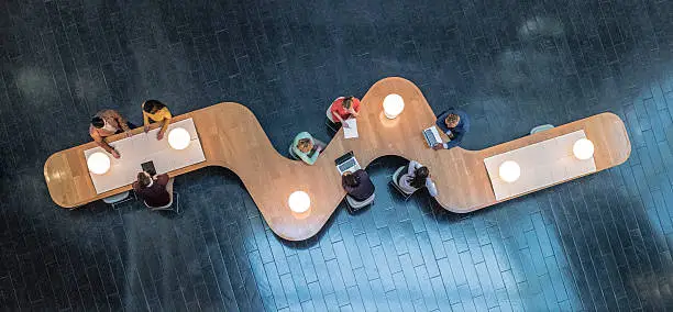 Panoramic overhead view of several business meetings going on in the communal area of a modern office building.