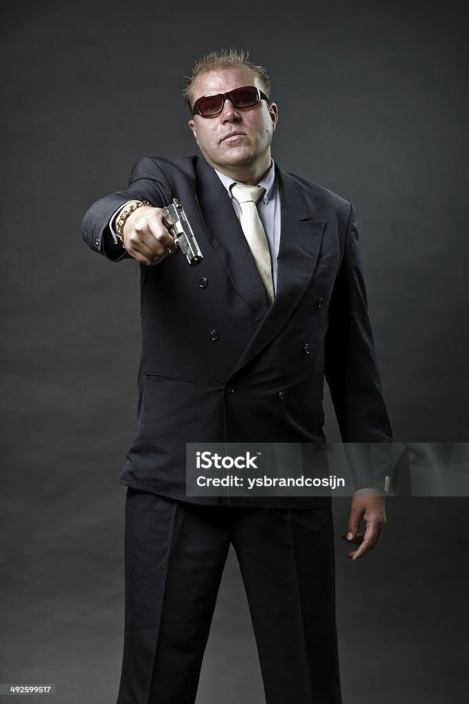 Gangster mafia man in suit with sunglasses holding a gun. Gangster mafia man in suit with tie and sunglasses holding a gun looking tough isolated on dark background. Adult Stock Photo