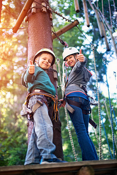 Brother and sister having fun in ropes course adventure park Little girl aged 9 with little brother aged 6, wearing helmets staning on wooden platform holding zip line in the outdoors ropes course adventure park. Kids are smiling at the camera showing thumbs up. canopy tour photos stock pictures, royalty-free photos & images