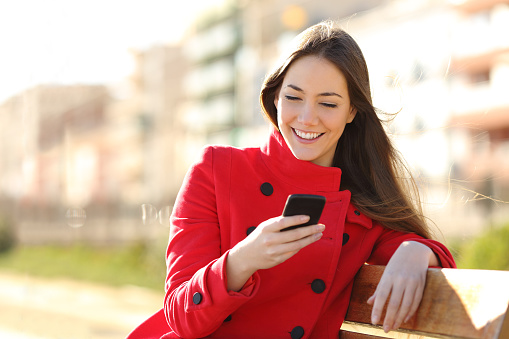 Girl texting on the smart phone sitting in a park wearing a red jacket and sitting in a bench in a park