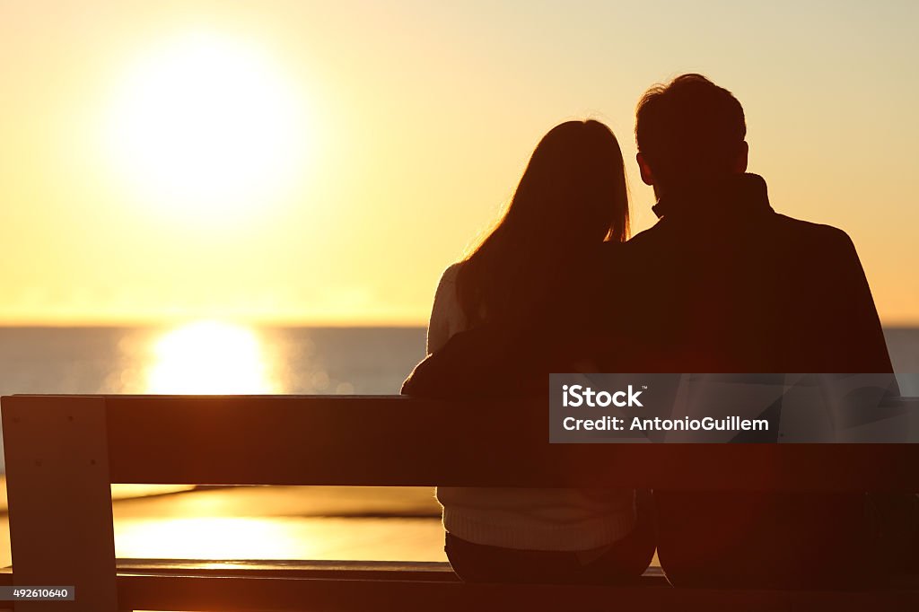 Back view of a couple watching sun on the beach Back view of a couple silhouette hugging and watching sun on the beach Couple - Relationship Stock Photo