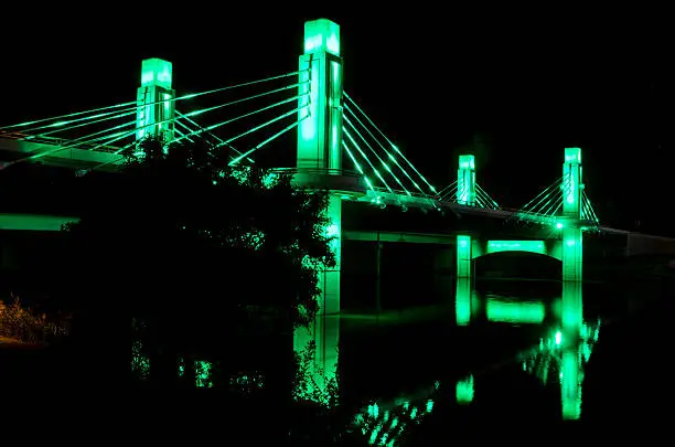 One of two pedestrian bridges leading to McLane Stadium lit up in the Baylor colors of green and gold is reflected in the Brazos River.  McLane Stadium is the brand new football venue on the campus of Baylor University.  It stands on the banks of the Brazos River, providing the latest in sports and technology for unversity students and supporters, as well as an area along the river to provide "sail gating," where sailboats can tie off and celebrate the pregame atmosphere.