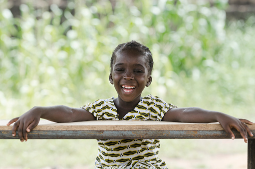 Gorgeous African schoolgirl laughing with a wonderful toothy smile sitting in her desk in an African school. Happiness fun symbol outdoors.