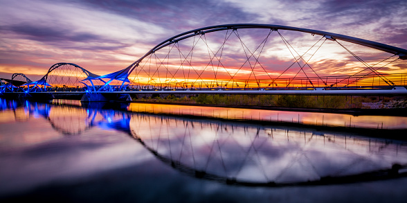 The sun is setting behind colorful clouds in the background of this picture of the walking bridge over Tempe Town Lake in Tempe, Arizona.