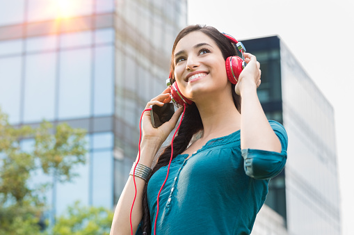 Closeup shot of young woman listening to music with mobile phone in the city center. Happy smiling girl listening to music with professional red headset. Beautiful brunette young woman feeling free and thinking.