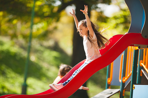 Little girl having fun in the park and sliding with her arms raised.