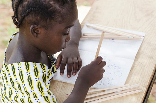 Back to school symbol: cute African black ethnicity girl drawing with lots of colorful pencils in her exercise book. Learning to draw activity outdoors. Beautiful young woman sitting in her desk.