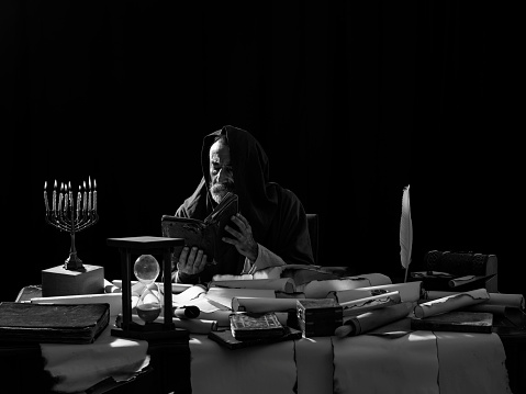Medieval philosopher reading Torah under the light of menorah.He is sitting on a desk.There are large amount of scrolls,books and box on desk.A menorah is placed on the left side of horizontal frame and a white quill pen on the right side.The image was shot with a medium format camera Hasselblad in color then edited to black and white.