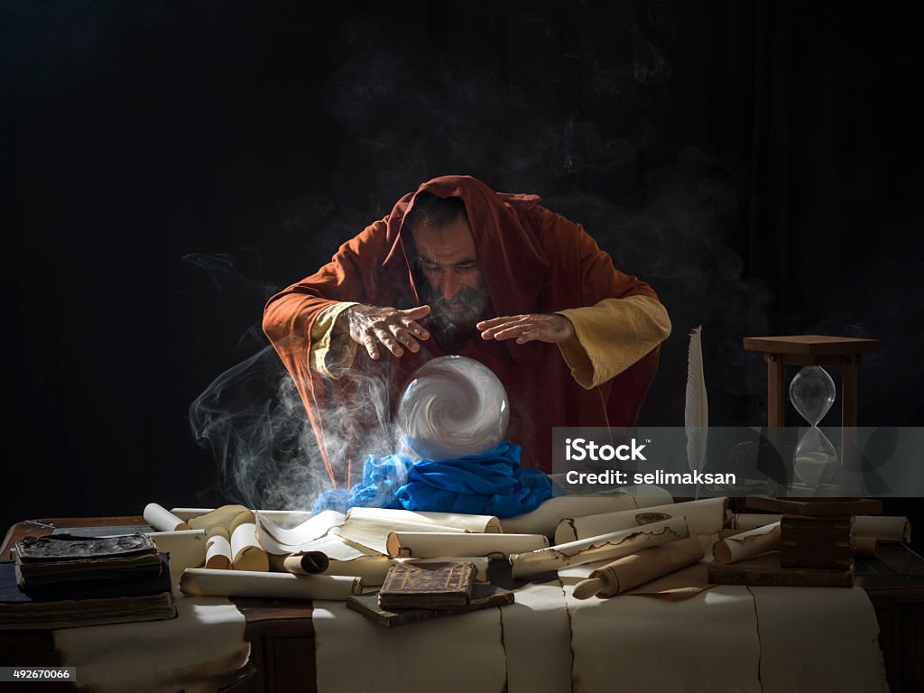 Fortune teller in fantastical costume using crystal ball Fortune teller magician in fantastical smoky atmosphere using crystal ball.He is wearing a brown frock with hood.The background is black.Smoke is coming out from the desk.There are large amount of scrolls, few antique black books and a hourglass on desk.Shot with a medium format camera Hasselblad. Wizard Stock Photo