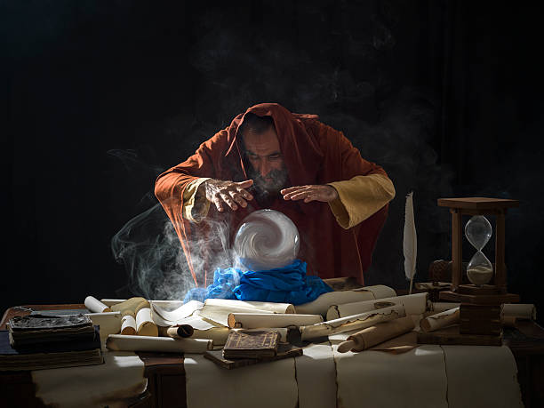 Fortune teller in fantastical costume using crystal ball Fortune teller magician in fantastical smoky atmosphere using crystal ball.He is wearing a brown frock with hood.The background is black.Smoke is coming out from the desk.There are large amount of scrolls, few antique black books and a hourglass on desk.Shot with a medium format camera Hasselblad. wizard stock pictures, royalty-free photos & images