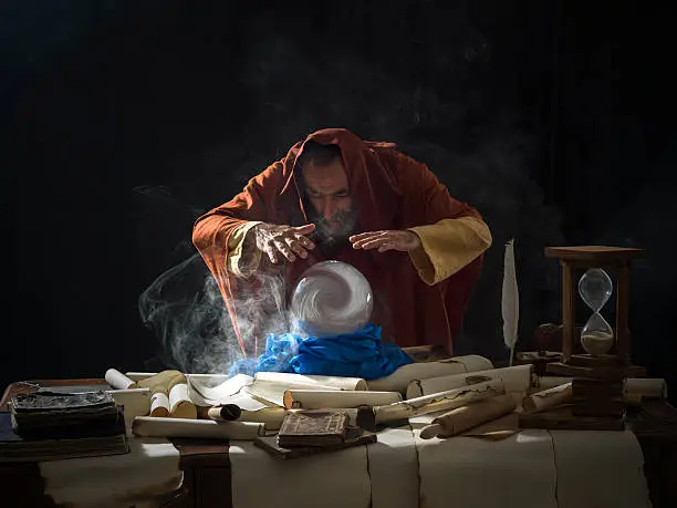 Fortune teller magician in fantastical smoky atmosphere using crystal ball.He is wearing a brown frock with hood.The background is black.Smoke is coming out from the desk.There are large amount of scrolls, few antique black books and a hourglass on desk.Shot with a medium format camera Hasselblad.