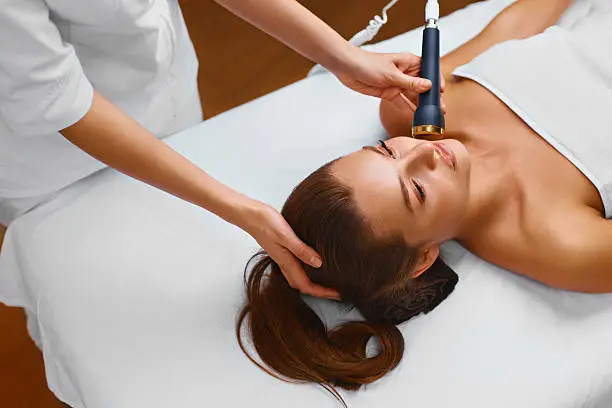 Face skin care. Beautiful young healthy caucasian woman lies on a table in a medical beauty cosmetology spa salon getting facial skin care treatment. Ultrasound cavitation anti-aging, rejuvenation, lifting procedure. Beautician applying regenerative, moisturizing cosmetics. Tightening of facial skin, restoration of youthful skin, anti-wrinkle. Beauty concept